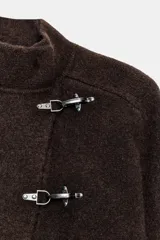 Chaqueta corta de punto marrón con cuello subido, manga larga y cierre frontal cruzado con alamares metálicos plateados.