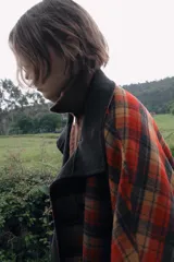 Capa oversize de corte amplio con estampado de cuadros en tonos rojos, negros y marrones. Presenta cuello alto, cierre frontal con botones, bolsillos delanteros y mangas ajustables mediante trabillas con botones.
