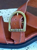 Sandalias de cuero color marrón con tiras anchas cruzadas y hebilla metálica decorativa en forma de estribo. Presentan taco cuadrado de altura media y suela de goma.