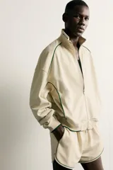 Chaqueta estilo deportivo de corte relajado, color beige con detalles de vivos en color verde oscuro. Presenta cuello alto, cierre frontal con cremallera de doble dirección, bolsillos laterales y puños elásticos.