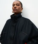 Chaqueta bomber de denim azul oscuro con cuello alto, cierre frontal con zíper y cintura ajustable con cordón. Presenta mangas largas con puños elásticos y bolsillos frontales funcionales.