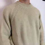 Sweater de tejido de punto grueso en color beige, con cuello redondo y mangas raglán. Presenta un diseño holgado y una pequeña etiqueta con logo en el puño.