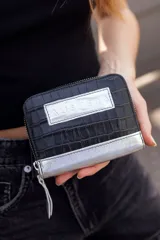 Billetera chica de cuero negro con textura tipo cocodrilo y detalles plateados. Cuenta con cierre y logo de la marca en el frente.