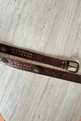 Cinturón de cuero vacuno color chocolate con hebilla metálica en tono oro viejo. Presenta apliques metálicos circulares grabados y tachas decorativas a lo largo de la tira.