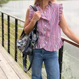 Camisa sin mangas de corte recto, con diseño a rayas verticales en tonos fucsia, azul y blanco. Presenta cuello alto, cierre frontal con botones y volados fruncidos a lo largo de la botonera y en las sisas.