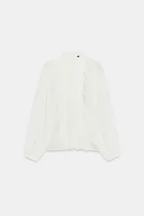 Blusa de bambula color blanco con cuello subido y manga larga. Presenta detalles de volantes y puntillas a tono en el frente, con cierre posterior mediante abertura y botón.