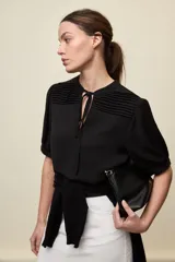 Blusa de manga corta color negro, con calce relajado, escote redondo con abertura y lazo para anudar. Presenta detalles de tablas horizontales en la parte superior que aportan textura al diseño.