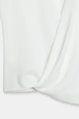 Top blanco de manga sisa con cuello redondo y diseño drapeado cruzado en la parte frontal, con bajo asimétrico y cierre en la espalda con abertura y botones.
