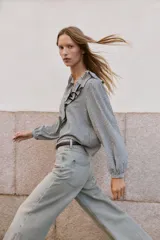 Blusa de manga larga con estampado de cuadros vichy en blanco y negro. Presenta un escote en V adornado con un volante amplio y un lazo decorativo en el centro. Las mangas terminan en puños elásticos.