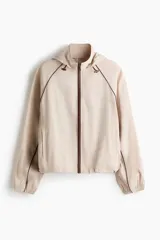 Campera rompevientos de color beige, modelo oversized con cuello alto y capucha con cordón ajustable. Cuenta con cierre frontal marrón, mangas raglán con puños elásticos, bolsillos laterales y detalles de ribetes en tono marrón oscuro.