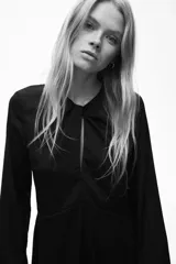 Vestido corto de color negro, con escote redondo y detalle girado en el frente. Presenta mangas largas con abertura en los puños, corte entallado en el torso y falda con leve vuelo. Cuenta con cierre oculto en la espalda.