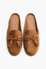 Mules de gamuza color marrón tostado, estilo mocasín, con puntera redondeada, detalle de lazo con borlas en el empeine y diseño destalonado.