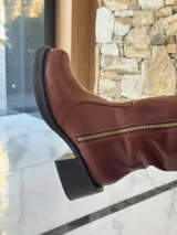 Bota de caña alta estilo biker, confeccionada en cuero color chocolate. Presenta un cierre metálico lateral que recorre toda la caña, estribo y cadena decorativa desmontables, y taco cuadrado con plataforma.