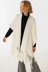 Ruana tejida color crudo, de punto grueso, con mangas cortas y terminación de flecos en el ruedo.