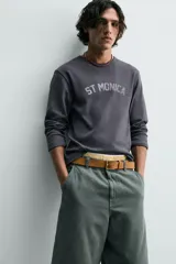 Camiseta de manga larga, corte ajustado (muscle fit), color gris oscuro con efecto lavado. Presenta un estampado frontal desgastado con el texto curvo "ST MONICA" en color plateado o gris claro.