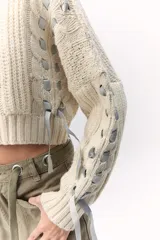 Sweater de lana merino tejido en punto grueso, color gris oscuro, con cuello alto. Presenta un diseño calado con tientos de cuero marrón insertados a lo largo de la prenda, simulando un lazo o detalle orgánico.