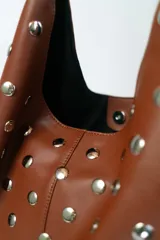 Bolso tipo hobo de cuero marrón, adornado con tachas metálicas redondas plateadas en toda su superficie. Posee un asa de hombro única y delgada.