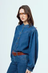 Camisa de denim azul oscuro con cuello mao, mangas abullonadas con puños elastizados y cierre frontal de botones metálicos.