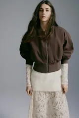 Campera bomber color marrón oscuro de tejido grueso, con cuello corto, puños y bajo acanalados. Cuenta con cierre frontal plateado, bolsillos frontales insertados y diseño de hombros caídos. Está completamente forrada.