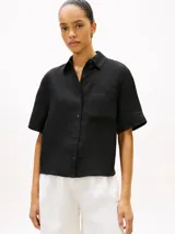 Camisa de manga corta de corte amplio confeccionada en puro lino, de color negro, con cuello inglés y un bolsillo de parche en el pecho que incluye un sutil logo bordado. Presenta hombros ligeramente caídos y cierre frontal de botones.