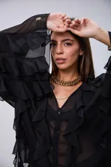 Blusa de tela semitransparente color negro, con diseño de volados verticales a lo largo del frente y en las mangas. Presenta cierre frontal con botones y cuello en V.