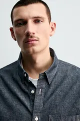 Camisa de corte relajado confeccionada en tejido de algodón denim, con cuello solapa, manga larga con puños abotonados y bolsillo plastrón en el pecho. Presenta cierre frontal de botones y bajo con aberturas laterales.