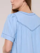 Blusa celeste de lyocell ultraliviano, de estilo romántico y silueta holgada. Presenta escote redondo con abertura en V y botones frontales, canesú fruncido y mangas cortas amplias.