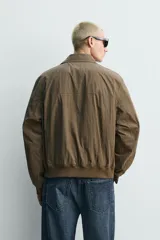 Cazadora regular fit de tejido técnico color marrón, con cuello solapa, manga larga y cierre frontal de cremallera. Presenta bolsillos de plastrón con solapa en la cadera y acabados con rib elástico.