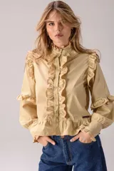Camisa celeste con volados decorativos en los hombros, pecho, mangas y ruedo, destacados con ribetes en tono rojo. Presenta cierre frontal de botones, cuello alto y mangas largas abullonadas con puños ajustados.