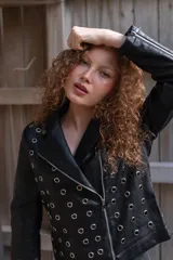 Campera de eco cuero negro con solapas anchas, cierre diagonal metálico y detalle de múltiples ojalillos metálicos distribuidos en el frente y las mangas.