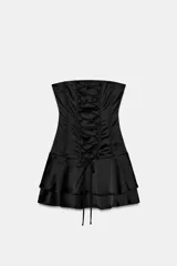 Vestido mini confeccionado en tejido satinado de color negro, con escote recto sin tirantes, detalles de lazos cruzados en el delantero estilo corsé y falda con volantes escalonados.