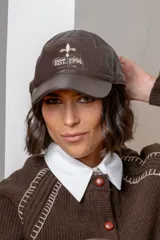 Gorra tipo baseball de cuero color chocolate, con bordado frontal en hilo beige que incluye una flor de lis y el texto 'EST 1994'.