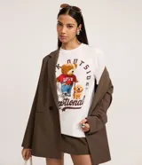 Remera blanca de algodón con cuello redondo y manga corta. Presenta un estampado grande de un oso de peluche con una camiseta roja que dice "LOVE" y un perro, rodeados de texto en color marrón.