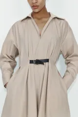 Vestido midi de color beige con cuello solapa y escote en pico. Presenta mangas largas, cintura elástica en la espalda y un cinturón a contraste con hebilla metálica. Incluye bolsillos laterales ocultos.