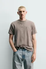 Remera de punto de algodón color gris topo, con efecto lavado, cuello redondo y manga corta. Cuenta con aplicación de parches con texto desgastado en el frente.