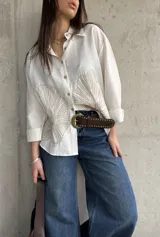 Camisa oversize blanca con botones frontales, cuello camisero y manga larga. Presenta grandes bordados en hilo beige con diseño de hojas o abanicos en la parte inferior frontal.