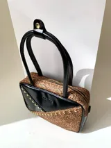 Cartera rígida negra con detalles en rojo y tachas plateadas. Tiene herrajes y cierre metálico plateado marca YKK, asa fina para llevar al hombro o en la mano y correa larga extra regulable. El interior es forrado, tiene un bolsillo con cierre enmarcado en cuero, y un segundo bolsillo plaqué.