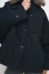 Trench corto de corte recto con capucha y cuello desmontable efecto pelo. Presenta cierre frontal con botones, bolsillos laterales y mangas largas con trabillas en los puños.