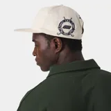 Gorra color beige con visera plana y parche bordado en el frente con escudo y la inscripción 'GOLF'.