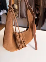 Cartera tipo hobo de textura gamuzada en color marrón oscuro, con diseño flexible y asa larga regulable. Presenta un detalle de flecos largos desmontables en uno de los laterales y cierre superior.