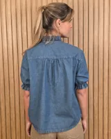 Camisa de denim de algodón celeste, con cierre frontal de botones, cuello alto con volados y mangas cortas abullonadas con puños elastizados fruncidos.
