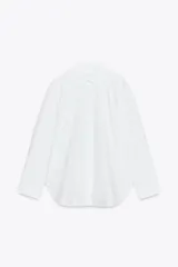 Camisa blanca de popelín de algodón 100%, con cuello solapa, manga larga terminada en puño y cierre frontal con botones ocultos por solapa. Presenta un corte holgado y ruedo curvo.