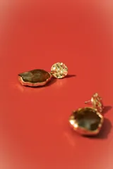 Aros colgantes dorados con forma irregular, engastados con una piedra o resina de color marrón oscuro y verde azulado veteado. El poste superior tiene un diseño orgánico y texturizado.