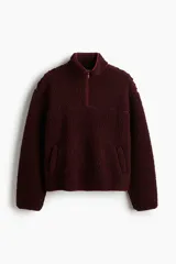 Buzo de sherpa con textura de borrego, color bordó, de corte oversized y hombros caídos. Presenta cuello elevado con medio cierre frontal, bolsillos laterales insertados y canesú en frente y espalda.