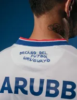 Camiseta de fútbol Umbro del Club Nacional de Football, modelo Home 2025, para hombre. Diseño blanco con detalles en azul y rojo en cuello y mangas. Incluye logo de Umbro, escudo del club y sponsor Antel en el frente.