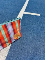 Sobre de mano rectangular con cierre superior y correa de muñeca, con diseño a rayas horizontales en tonos naranja, rojo, verde y celeste.