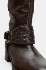 Bota de caña alta de cuero color marrón oscuro, estilo motociclista, con taco bajo y punta redonda. Presenta una tira con hebilla ajustable en la parte superior de la caña y una tira desmontable con argolla metálica y detalle de rosa repujada en el tobillo.