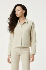 Chaqueta corta de algodón color beige claro, estilo chore coat o de trabajo, con cuello camisero, cierre frontal de botones y dos bolsillos grandes de parche en la parte delantera.