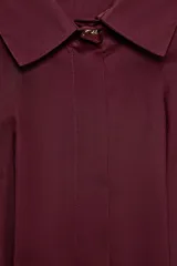 Camisa de manga larga color vino tinto, con cuello solapa y cierre frontal de botones ocultos. Presenta un diseño entallado en la cintura con pliegues verticales que generan un efecto peplum en el bajo.