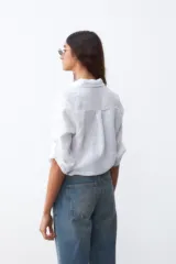 Camisa blanca de lino con cuello clásico, mangas 3/4 con botón para enrollar, bolsillo en el pecho y cierre frontal con botones. Se ata en la cintura.